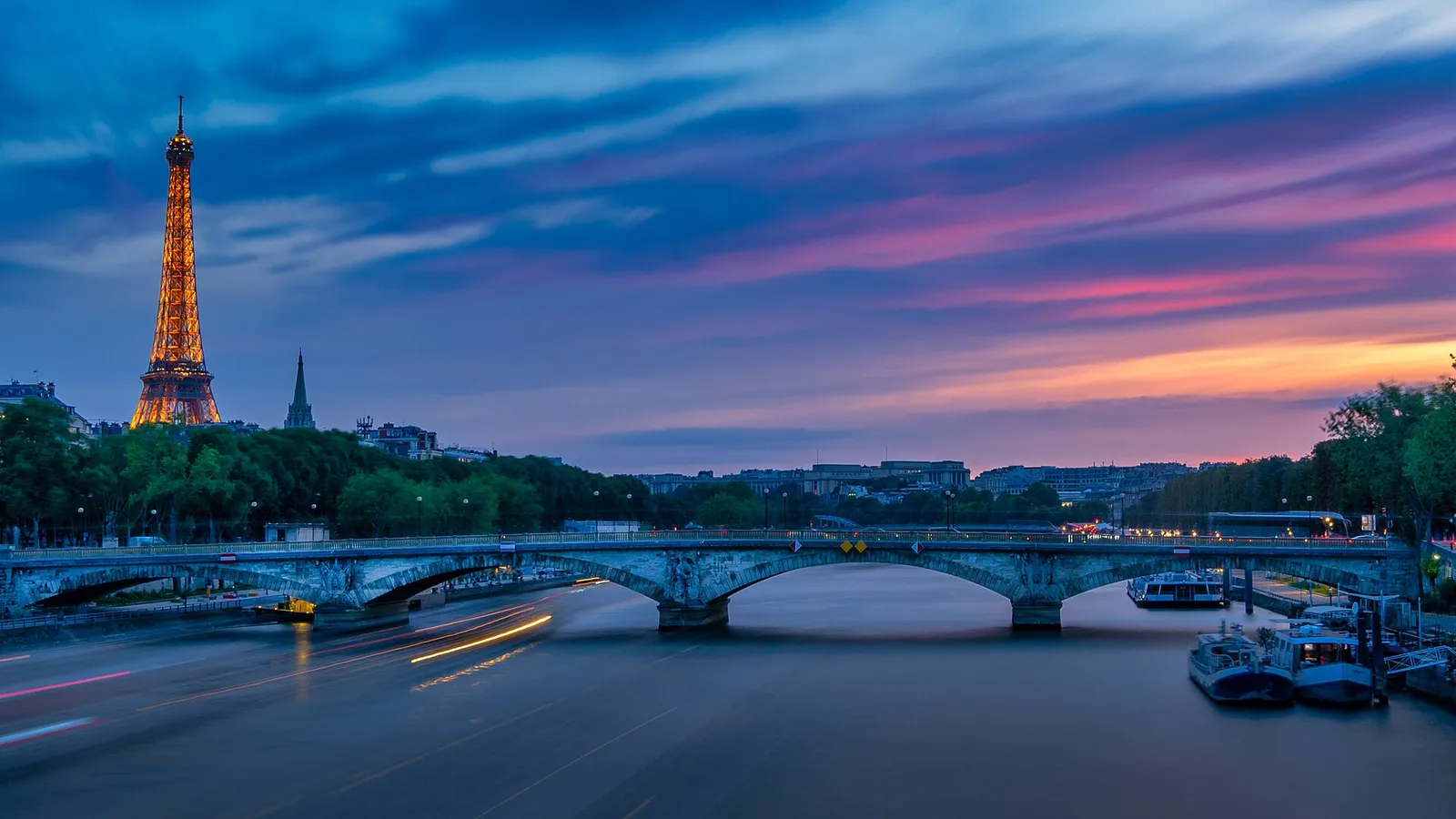 Eiffel Tower lit against a pink and blue sky behind the Pont des Invalides, with river boats moored at right, Seine near the Alma, sunset